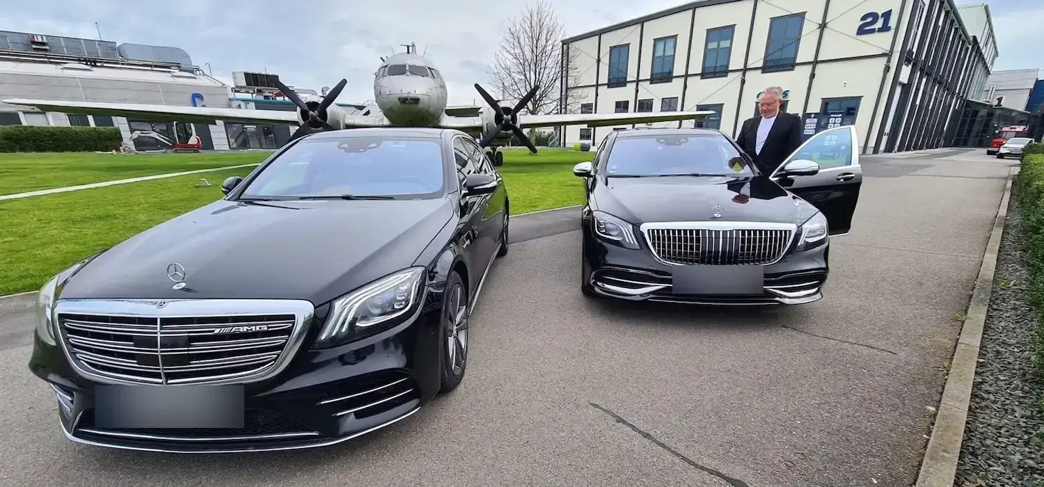 Chauffeur neben einer Mercedes-Maybach S-Klasse, links eine Mercedes S-Klasse, im Hintergrund ein historisches Flugzeug